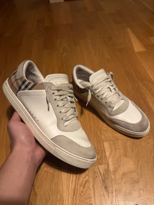 Burberry vita låga sneakers - Snygga vita sneakers från Burberry med klassiskt rutigt mönster på hälen, grå mockadetaljer och vita skosnören. Skorna har en låg profil och är tillverkade i en mix av läder och mocka. Perfekta för dig som vill ha en clean och stilren look med en touch av lyx.