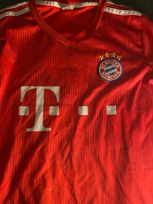 Bayern München matchtröja Lewandowski - Röd Bayern München matchtröja från Adidas med vita detaljer, klubbmärke på bröstet och sponsorlogga framtill. Nummer 9 och Lewandowski tryckt på ryggen. Klassisk V-ringning och tre ränder på axlarna. Perfekt för fotbollsfans!