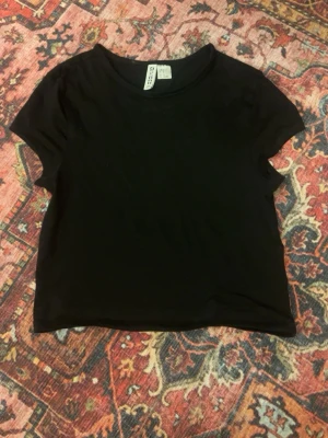 Svart cropped t-shirt från H&M - En enkel svart cropped t-shirt från Gina Tricot med rund halsringning och korta ärmar. Perfekt att matcha med höga jeans eller kjol. Tunn och mjuk bomullskvalitet som känns skön mot huden.