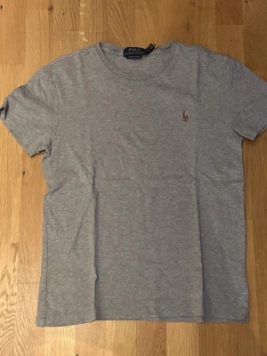 Grå t-shirt från Polo Ralph Lauren - Klassisk grå t-shirt från Polo Ralph Lauren med rund halsringning och broderad logga på bröstet. T-shirten är kortärmad och har en enkel, stilren design som passar till det mesta. Tillverkad i mjuk bomull för en skön känsla.