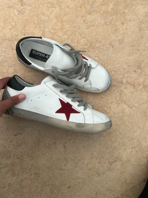Golden Goose vita sneakers med röd stjärna - Snygga vita sneakers från Golden Goose med grå skosnören, svart häl och en ikonisk röd stjärna på sidan. Skorna har en klassisk låg modell och är tillverkade i läder med slitna detaljer för en cool look.