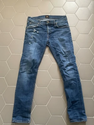 Blå slitna jeans från Lee - Snygga blå jeans från Lee med smal passform och slitna detaljer på framsidan. Klassisk femficksmodell med dragkedja och knapp. Jeansen har en cool tvättad look och Lee-lapp bak i midjan. Perfekta för en avslappnad och trendig stil.