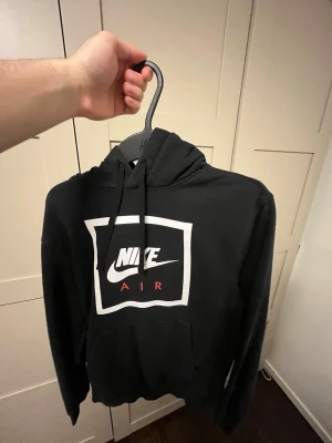 Svart Nike Air hoodie med tryck - Svart hoodie från Nike med stor vit och röd Nike Air-logga på bröstet. Klassisk huva med dragsnören och magficka framtill. Perfekt för en sportig och avslappnad stil.