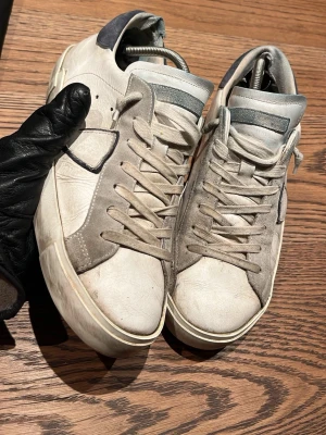 Vita Philippe Model sneakers - Snygga vita sneakers från Philippe Model med grå och blå detaljer. Skorna har en låg silhuett, klassisk snörning och är tillverkade i läder med mockadetaljer på sidan. Perfekta för dig som gillar stilrena och trendiga skor.