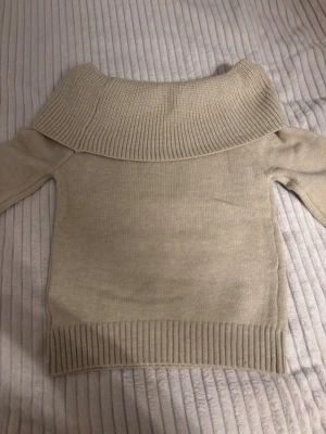 Beige off shoulder stickad tröja - En beige stickad tröja med off shoulder-krage och långa ärmar. Tröjan har ribbstickade muddar vid ärmslut och nederkant, vilket ger en snygg och bekväm passform. Perfekt för dig som gillar en stilren och trendig look. Ärmarna är lite utsvängda. Aldrig använd eftersom den inte passade så bra.