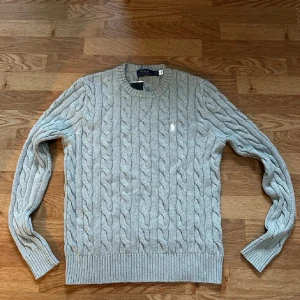 Grå kabelstickad tröja Polo Ralph Lauren - Säljer en grå kabelstickad tröja från Polo Ralph Lauren med klassisk rund hals och långärmad passform. Tröjan har det ikoniska vita Polo-logot broderat på bröstet och är tillverkad i mjuk bomull. Perfekt för dig som gillar stilren och tidlös design.