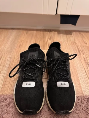 Svarta Adidas NMD sneakers - Säljer ett par svarta Adidas NMD sneakers med vit sula och svarta/vita skosnören. Ovandelen är i meshmaterial och har detaljer med texten 'NMD' framtill samt Adidas-logga på plösen. Bak finns textdetaljer på hälen. Sköna och stilrena sneakers.