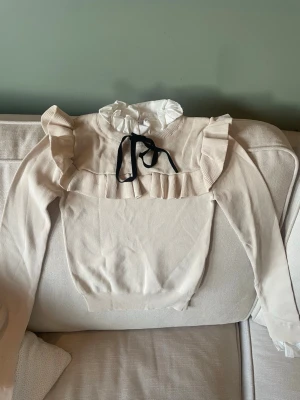 Beige volangblus med rosett från Zara - Supersöt beige blus från Zara med volangdetaljer över axlar och bröst, hög vit krage och svart rosett framtill. Långärmad modell i mjukt material, perfekt för en trendig och chic look.