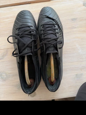 Svarta fotbollsskor i läder - Säljer ett par klassiska svarta fotbollsskor med snörning och rundad tå. Skorna har en stilren design med diskreta sömmar och är tillverkade i slitstarkt lädermaterial. Perfekta för dig som vill ha en tidlös look på planen. Använda cirka 1,5 månad på konstgräs, 3 dagar i veckan.  