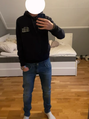 Mörkblå hoodie från Les Deux - Snygg mörkblå hoodie från Les Deux med vit logga på bröstet. Klassisk modell med huva och känguruficka framtill. Perfekt för en avslappnad och stilren look. Mjuk bomullsblandning och ribbade muddar vid ärmslut och nederkant. Passar dig mellan 170-180