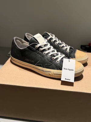 Golden Goose V-Star - Snygga svarta sneakers från Golden Goose med beige tåparti och sula. Skorna har vita snören, stjärndetalj på sidan och GGDB-text i guld. Ovandelen är i läder med slitna detaljer för en cool vintage-look. Perfekta för dig som gillar streetstyle.