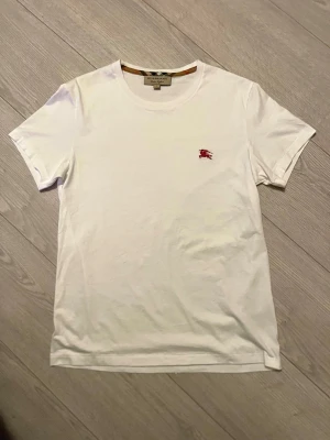 Vit t-shirt från Burberry - Klassisk vit t-shirt från Burberry med rund halsringning och det ikoniska röda Burberry-emblemet broderat på bröstet. Tillverkad i mjuk bomull och har en enkel, stilren design som passar till allt.