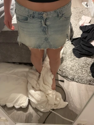 Ljusblå jeanskjol med rå kant - Trendig ljusblå jeanskjol med rå, fransig kant och snygga detaljer på bakfickorna. Kjolen har klassisk femficksdesign, knappgylf och är i en kort modell. Perfekt för dig som gillar en avslappnad och cool stil.