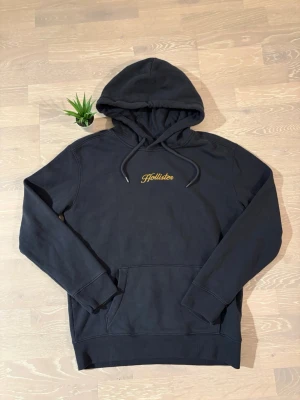 Svart hoodie från Hollister - Svart hoodie från Hollister med gul broderad logga på bröstet. Klassisk känguruficka framtill och justerbar huva med dragsko. Tillverkad i mjukt bomullsmaterial som är perfekt för chill dagar. Ribbade muddar vid ärmslut och nederkant.