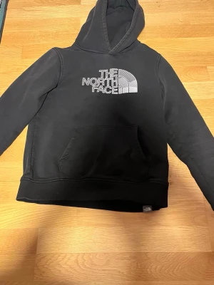 Svart hoodie från The North Face - Svart hoodie från The North Face med vit logga på bröstet. Klassisk känguruficka framtill och huva. Mjuk bomullsblandning som är skön att ha på sig. Perfekt för en avslappnad och cool stil.