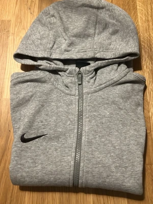 Grå zip-hoodie från Nike - Snygg grå hoodie från Nike med dragkedja framtill och svart Nike-logga på bröstet. Tröjan har huva och två fickor framtill. Tillverkad i mjukt bomullsmaterial, perfekt för en avslappnad stil.