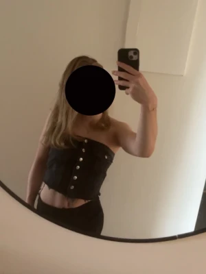 Svart jeans tubtopp med knappar - Säljer en svart tubtopp i jeansmaterial med silverfärgade knappar framtill och rå kant nedtill. Toppen har två dekorativa bröstfickor och en croppad passform. Perfekt för dig som vill ha en edgy och trendig look.