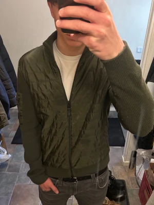 J.Lindeberg Cardigan Jacka - Snygg olivgrön cardigan jacka med quiltad front och rygg samt stickade ärmar och muddar. Dragkedja framtill och två fickor med dragkedja. Hög krage och patch på ena ärmen ger en modern look. Perfekt för lager-på-lager och streetwear.
