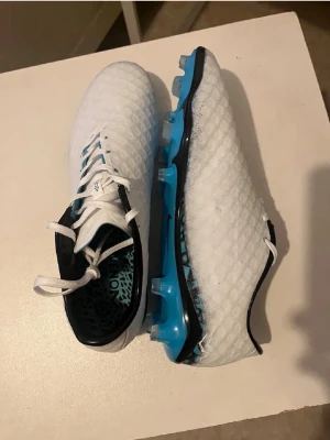 Vita Nike fotbollsskor med blå sula - Säljer ett par vita Nike fotbollsskor med blå detaljer och svart kant. Skorna har en texturerad ovansida för bättre bollkontroll och blå dobbar för bra grepp på planen. Snygg design med snörning och mönstrad innersula. Perfekta för dig som vill sticka ut på fotbollsplanen.