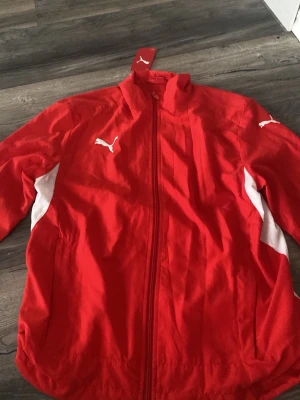 Röd träningsjacka från Puma med prislappen kvar - Snygg röd träningsjacka från Puma med vita detaljer på sidorna och ärmarna. Jackan har dragkedja framtill, ståkrage och elastiska muddar vid ärmsluten. Perfekt för träning eller sportiga outfits. Puma-logga på bröstet och ärmen. Pris lapp kvar!!!
