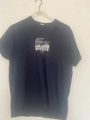 Mörkblå Lacoste t-shirt med tryck - Snygg mörkblå t-shirt från Lacoste med klassiskt krokodiltryck och text på bröstet. T-shirten har rund halsringning och korta ärmar. Perfekt för en stilren och sportig look.