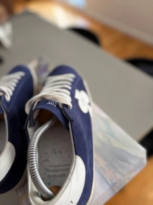 Blåa The Gallery sneakers med vit detalj - Nytt skick som kommer med original box.  Snygga sneakers från The Gallery i blå mocka med vita detaljer och vit sula. Skorna har vita skosnören och en vit blomdetalj på sidan. Perfekta för dig som gillar en stilren och trendig look.