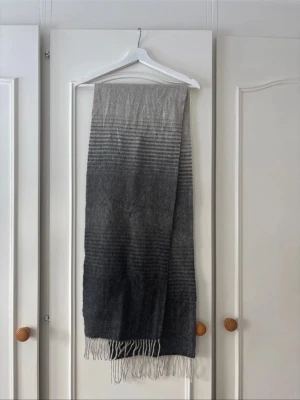 Gråmelerad halsduk med fransar - Snygg och lång halsduk i gråmelerad ton med ränder och fransar i ändarna. Halsduken är 100% ull och är ca 183 cm lång exklusive fransarna. Mjuk och skön, perfekt till höst, vår och vinter. Passar till de flesta outfits och ger en stilren look.