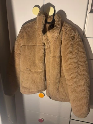 Beige teddyjacka från New Look - Supermjuk och fluffig teddyjacka i beige från New Look. Jackan har hög krage, dragkedja framtill och två fickor. Perfekt för kalla dagar när du vill vara både varm och snygg. Materialet är fuskpäls som ger en mysig känsla.