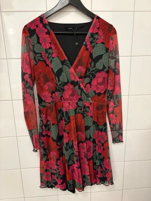 Blommig omlottklänning från Bik Bok - Snygg kort omlottklänning från Bik Bok med blommigt mönster i rött, rosa och grönt på svart botten. Klänningen har v-ringning, långa ärmar i mesh och volangkant nertill. Perfekt för dig som vill sticka ut med färg och mönster.