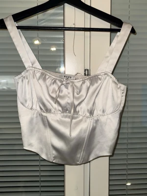 Vit satin korsett från NA-KD - Snygg vit korsettopp från NA-KD i glansigt satinmaterial. Toppen har breda axelband, croppad passform och en söt knytdetalj framtill. Perfekt att styla med jeans eller kjol för en trendig look.