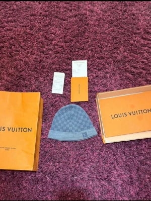 Louis vuitton petit damier mössa - Louis vuitton petit damier mössa i färgen ljusblå. Kommer med kvitto, låda och påsen. Lådan är skadad annars är allt annat i bra skick. Mössan är lite skadad på bakre delen där den är sydd men det är inget man märker när mössan är på