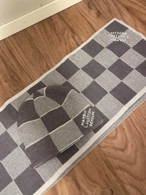 Louis Vuitton grå rutig halsduk - Grå och vit halsduk från Louis Vuitton med klassiskt rutigt mönster och broderad text 'Marque Louis Vuitton Déposée' i ena hörnet. Halsduken har en rektangulär form och är tillverkad i ett mjukt stickat material. Perfekt för att ge din outfit en exklusiv touch.