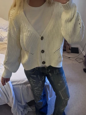 Stickad kofta - Säljer en krämvit stickad kofta med stora bruna knappar och v-ringning. Koftan har ett chunky, ribbstickat mönster och ballongärmar. Perfekt att slänga över en t-shirt för en avslappnad look. Från Nakd
