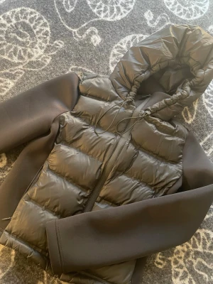 Svart pufferjacka med huva - Snygg svart pufferjacka med glansig vadderad front och huva med dragsko. Ärmar och sidor är i ett mjukare, matt tyg som ger en cool kontrast. Jackan har dragkedja framtill och en modern, avslappnad passform. Perfekt för kalla vinterdagar.