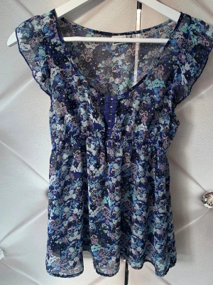 Blommig blå transparent blus - Söt blommig blus i blå toner med transparent material och volangärmar. Blusen har knappar framtill och ett lätt utställd passform. Perfekt för dig som gillar en luftig och somrig stil. Materialet känns tunt och flowy.