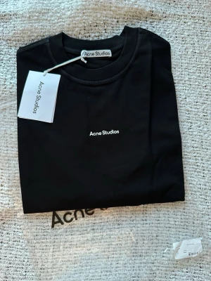 Svart Acne S. t-shirt - Svart t-shirt från Acne Studios med vit logga tryckt på bröstet. Klassisk rund halsringning och korta ärmar. Tillverkad i mjuk bomull för en clean och stilren look. Perfekt för dig som gillar minimalistisk design.