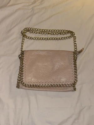 Beige tiamo axelväska - Beige axelväska i skinn med guldfärgad kedja som axelrem och detaljer runt kanterna. Väskan har ett fack på baksidan och lock framtill. Jag har aldrig använt den. SKRIV FÖR FRAKT💞