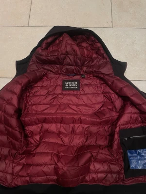 Jacka från Scotch & Soda S - Säljer en svart pufferjacka från Scotch & Soda i storlek S. Jackan har vinrött quiltat foder, huva och två snedställda fickor med dragkedja. Perfekt för kalla dagar och har en clean look med diskreta detaljer.