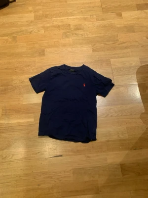 Mörkblå t-shirt från Polo Ralph Lauren - Mörkblå t-shirt från Polo Ralph Lauren med klassisk passform och korta ärmar. T-shirten har en liten röd broderad logga på bröstet och är tillverkad i mjuk bomull. Perfekt för en stilren och avslappnad look.