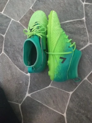 Grönblå Adidas fotbollsskor - Säljer ett par grymma fotbollsskor från Adidas i en snygg grön och blå färgkombination. Skorna har högre skaft, snörning och klassiska svarta Adidas-ränder på sidan. Ovandelen har textur för bättre bollkontroll och yttersulan är gjord för bra grepp på planen.