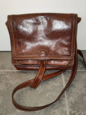 Brun axelväska i läder från The Bridge - Snygg brun axelväska i äkta läder från The Bridge. Klassisk rektangulär form med lock och justerbar axelrem. Väskan har dekorativa sömmar och en liten metallogo framtill. Perfekt för dig som gillar vintagevibbar och tidlös design.