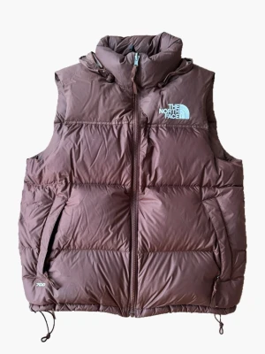 Brun dunväst från The North Face - Snygg brun dunväst från The North Face med hög krage och dragkedja framtill. Västen har två sidofickor med dragkedja och broderad logga på bröstet. Tillverkad i slitstarkt material och fylld med dun för extra värme. Perfekt för lager-på-lager under kalla dagar.