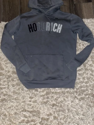 Grå HOODRICH hoodie med tryck - Snygg grå hoodie från HOODRICH med stor magficka och huva med snörning. Framtill står det HOODRICH i svart, grått och vitt tryck. Hoodien har långa ärmar och ribbade muddar. Perfekt för en chill och street-inspirerad look.