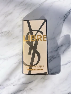 Yves Saint Laurent Libre EdP 90ml - Yves Saint Laurent Libre Eau de Parfum 90ml i elegant beige och svart förpackning med guldiga detaljer. Doften är modern och lyxig, perfekt för dig som gillar exklusiva parfymer. Kartongen har en stilren design med stora YSL-bokstäver och guldtext.