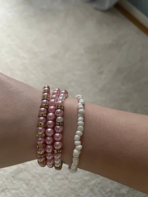 Set med pärlarmband i rosa och vitt - Säljer ett set med fyra elastiska armband med pärlor. Tre av armbanden har glansiga rosa pärlor och guldfärgade detaljer, medan det fjärde har små vita pärlor. Perfekt för dig som gillar att mixa och matcha accessoarer.