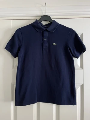 Mörkblå piké från Lacoste - En riktigt fin mörkblå pikétröja från Lacoste med korta ärmar, knappar och krage. 