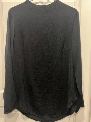 Svart långärmad blus med struktur från H&M - En svart blus med lång ärm och lätt strukturerat tyg. Modellen har rundad halsringning och något lös passform. Materialet ser ut att vara mjukt och stretchigt, perfekt för en clean och enkel look. Inga synliga detaljer eller mönster förutom den diskreta strukturen i tyget.