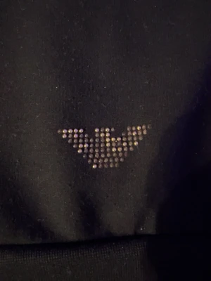 Svart hoodie från EA7 Emporio Armani - Snygg svart hoodie med dragkedja från EA7 Emporio Armani. Märket syns med glittrande stenar på bröstet och logga i samma stil. Hoodien har huva med dragsko och ribbade muddar. Perfekt för en trendig och avslappnad look.