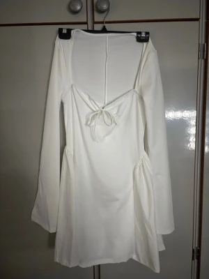 Vit kortklänning med knyt och v-ringning - Säljer en stilren vit kortklänning med lång ärm och v-ringning framtill. Klänningen har en snygg knyt-detalj vid bysten och lätt utsvängda ärmar. Perfekt för dig som gillar minimalistisk och trendig stil.