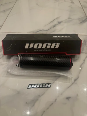 VOCA kolfiber ljuddämpare - Säljer en VOCA ljuddämpare i svart kolfiber med röda detaljer. Cylindrisk form och kommer med originalkartong samt VOCA-dekal. Perfekt för dig som vill uppgradera ljudet och stilen på din moped eller motorcykel.